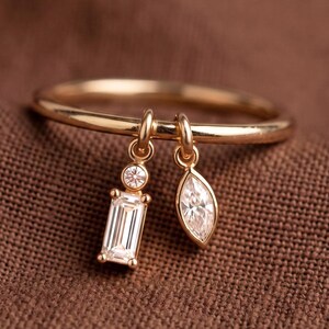 Op de afbeelding: Een gouden ring met twee bungelende bedels. De ene bedel is een rechthoekige baguette-geslepen heldere steen met een kleine ronde steen erboven, de andere bedel is een markies-geslepen heldere steen. De ringband is dun en gepolijst.