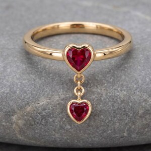 May include: A gold ring featuring a heart-shaped red gemstone set into the band, with a second, smaller heart-shaped red gemstone dangling from a short gold chain link attached directly below the primary heart setting.
