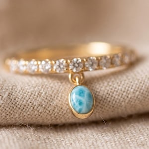 Könnte beinhalten: Ein goldener Ring mit einer Ringschiene, die mit einer Reihe kleiner, klarer, runder Steine besetzt ist, und einem einzelnen ovalen, hellblauen Steinanhänger, der in der Mitte hängt. Das Metall hat ein poliertes Finish und ein feines Glied.