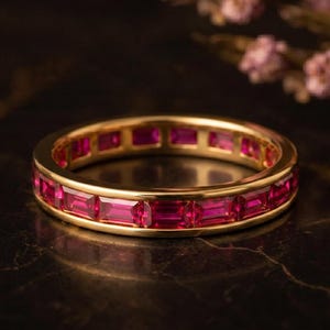 Puede incluir: Una alianza de oro con una hilera continua de vibrantes rubíes de color rojo rosado, tallados en forma rectangular y engastados en un canal de oro amarillo pulido. El anillo presenta un diseño uniforme con piedras en todo el contorno.