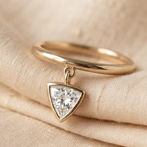 Peut inclure: Une bague en or avec une breloque pendante en forme de triangle. La breloque est sertie d'une pierre triangulaire transparente et facettée qui reflète la lumière. L'anneau de la bague est en or lisse et poli, et la breloque est fixée par une petite boucle.