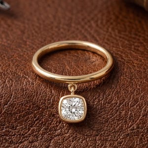 May include: A gold ring featuring a thin, polished band and a single dangling charm. The charm is a square-shaped, cushion-cut clear gemstone set in a gold bezel, which hangs from a small, circular jump ring attached to the main band.