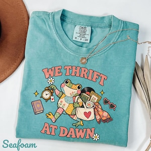 May include: A seafoam green t-shirt featuring a graphic of a patchwork frog carrying a tote bag, a clock, and books. The text reads "WE THRIFT AT DAWN" in pink letters. The shirt has a crew neck and a Comfort Colors brand label on the inside collar.