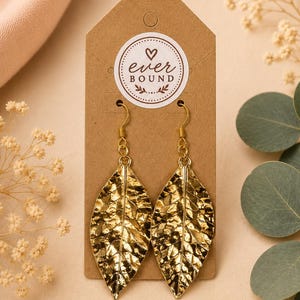 May include: A pair of gold-toned leaf-shaped earrings with a textured, metallic finish, suspended from gold-colored hooks. The earrings are attached to a rectangular brown cardboard display card featuring a circular logo that reads ever BOUND.