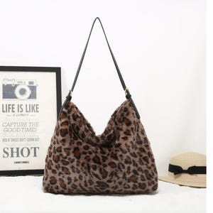 May include: A hobo-style shoulder bag featuring a brown and black leopard print faux fur exterior. The bag has a single thin black strap attached with gold-tone hardware, creating a slouchy silhouette with a wide opening at the top.