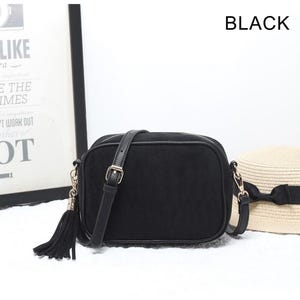 May include: A black rectangular crossbody bag with a smooth texture, featuring a long adjustable shoulder strap, a gold-toned metal buckle, and a matching black tassel attached to the side hardware. The word BLACK is printed in the upper right corner.
