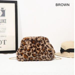 May include: A brown leopard print faux fur clutch bag with a gathered top closure and a thin gold-tone metal chain shoulder strap. The bag features a soft, textured surface with dark brown and black spots on a tan background.