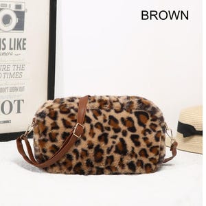 May include: A rectangular crossbody bag featuring a soft, faux fur exterior with a brown and black leopard print pattern. The bag includes a thin, adjustable brown strap made of a smooth material and gold-toned metal hardware clips on each side.