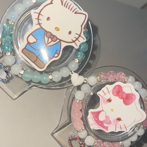 Può includere: Due braccialetti di perline con ciondoli di Hello Kitty. Un braccialetto ha perline azzurre e trasparenti con distanziatori argentati, mentre l'altro ha perline rosa e bianche con distanziatori argentati. Entrambi includono un ciondolo a forma di cuore.