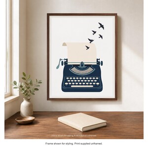 May include: A framed wall art print featuring a dark blue vintage typewriter with a blank sheet of paper emerging from the top. Four small black birds are depicted flying upwards from the paper against a plain white background.