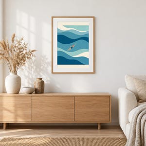 May include: A framed wall art print featuring a stylized illustration of a person swimming in ocean waves of varying shades of blue and white. The swimmer wears a dark blue swimsuit and an orange swim cap, centered within the wavy composition.
