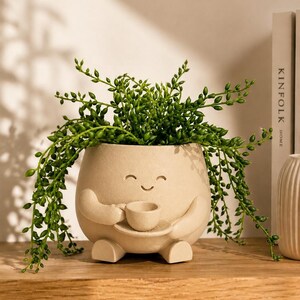 May include: A beige ceramic planter shaped like a round character with arms and legs, holding a small cup and saucer. The planter features a simple carved smiling face and is filled with a trailing green string of pearls succulent plant.