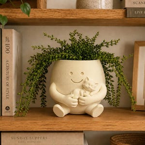 Op de afbeelding: Een crèmekleurige keramische plantenpot in de vorm van een zittend figuur met een lachend gezicht, die een kleine slapende kat in de armen houdt. De pot bevat een hangende groene plant met kleine ronde blaadjes op een houten plank.
