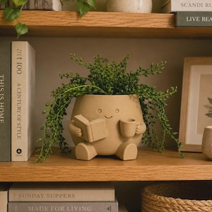 Puede incluir: Una maceta de cerámica de color tostado mate con forma de figura sentada con cara sonriente, que sostiene un libro abierto en una mano y una taza pequeña en la otra. La maceta contiene una planta verde colgante con pequeñas hojas redondas.