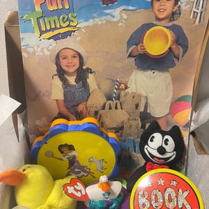 May include: A collection of vintage McDonald's toys including a Fun Times box, a yellow plush duck with a Ty tag, a blue and yellow tambourine featuring a character, a small plastic figure, a black plush cat, and a round red and yellow Book button.