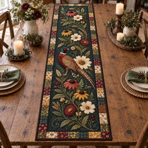 May include: A rectangular table runner featuring a dark green central panel with a printed design of a pheasant surrounded by various flowers and foliage, bordered by a patchwork pattern of small squares in shades of red, gold, and green.