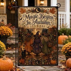 以下が含まれることがあります： Magic Begins In Autumnという文字が書かれた長方形のガーデンフラッグ。魔女の帽子とマントを身に着けた3人の人物、黒猫、カボチャ、キノコ、そしてオレンジ、茶色、濃い緑色の秋の葉が描かれています。