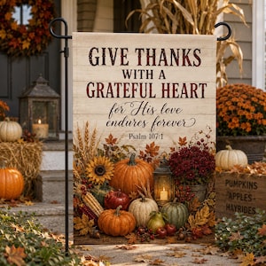 May include: A rectangular garden flag with a wood-plank background featuring the text "GIVE THANKS WITH A GRATEFUL HEART for His love endures forever Psalm 107:1" above an arrangement of pumpkins, sunflowers, wheat, berries, and a lantern.