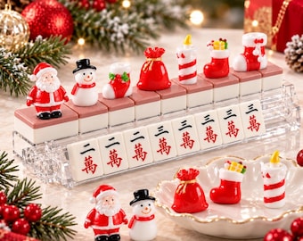 以下が含まれることがあります： サンタクロース、雪だるま、靴下、ギフトバッグ、キャンドルのミニチュアフィギュアが付いたクリスマス仕様の麻雀牌セット。透明なアクリル製のディスプレイスタンドに並べられており、牌には赤いインクで中国語の文字が印刷されています。