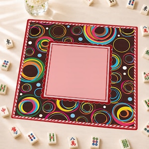 May include: A square game mat featuring a dark brown background with colorful concentric circles in shades of blue, yellow, pink, and green. A solid light pink square with a scalloped red border sits in the center, surrounded by mahjong tiles.
