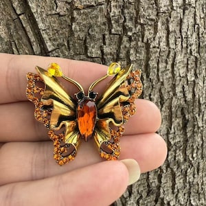 Puede incluir: Un broche de mariposa con acabado dorado metálico, una gran piedra de color ámbar en forma de lágrima en el centro, dos pequeños ojos circulares negros y alas decoradas con numerosos cristales pequeños de color naranja y dos piedras amarillas.