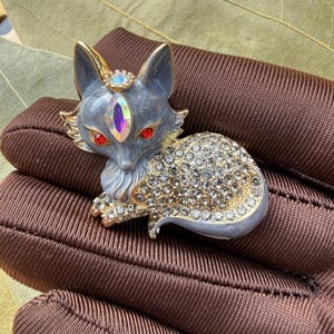 Puede incluir: Un broche con forma de zorro que presenta un acabado de esmalte gris con detalles en metal dorado. El zorro tiene dos ojos de cristal rojo, un cristal iridiscente de corte marquesa en la frente y un pequeño cristal redondo entre las orejas. Su cuerpo está cubierto de pedrería transparente.