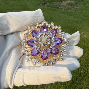 Puede incluir: Un broche circular con una base de metal dorado, una piedra central iridiscente rodeada por un anillo de cristales ovalados de color púrpura, acentuado por varias capas de pequeños diamantes de imitación redondos y claros.