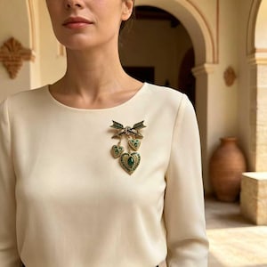 Puede incluir: Un broche de tono dorado con un diseño de lazo, dos flechas cruzadas y tres colgantes en forma de corazón. Cada corazón está acentuado con una piedra verde central y detalles de filigrana, prendido en una blusa de manga larga color crema.