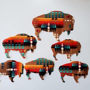 May include: Five fabric cutouts shaped like bison, each featuring a vibrant geometric pattern in shades of orange, red, turquoise, black, and white. The designs incorporate repeating triangles and diamonds across the body of each animal figure.