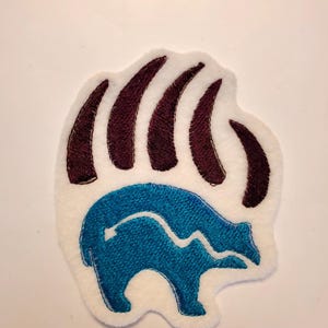 May include: An embroidered patch on a white felt backing featuring a dark brown bear paw print design. Inside the paw print, there is a stylized blue bear silhouette with a white line running across its body, creating a distinct layered graphic.