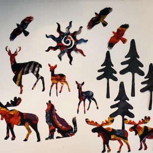 May include: A collection of flat, cutout shapes featuring a sun, three moose, two deer, one fawn, a howling wolf, three birds in flight, and four pine trees. The animal and sun shapes have a multicolored, patterned finish, while the trees are solid black.