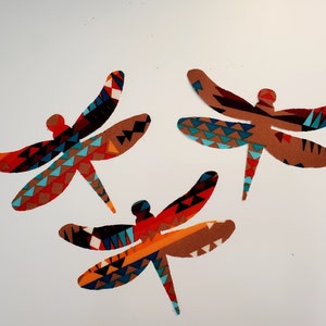 May include: Three decorative dragonfly cutouts with patterned wings in shades of brown, red, orange, blue, and black. The dragonflies are arranged on a white background. The wings feature geometric designs.