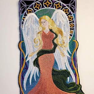 May include: Embroidered patch featuring an angel with flowing blonde hair, wearing a peach-colored gown and white wings. The angel is surrounded by a decorative frame with gold and purple accents.