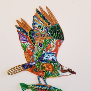 May include: A colorful illustration of an osprey in flight, carrying a fish. The bird's wings and body are adorned with intricate patterns in shades of orange, blue, green, and yellow. The fish is green and blue.