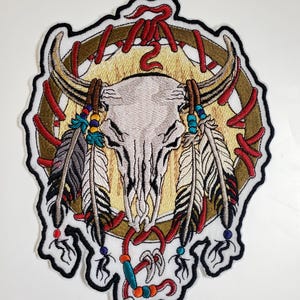 May include: Embroidered patch featuring a cream-colored bull skull with horns, adorned with feathers and beads. The design includes a gold-colored circle with red accents and a black border. A decorative item.