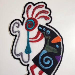 May include: Embroidered patch featuring a colorful Native American Kachina doll design. The doll is depicted in a traditional dance pose with a headdress and intricate details. The patch is made of felt and has a black outline.