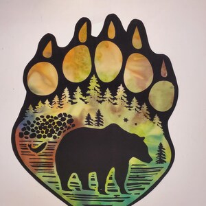 May include: A bear paw-shaped cutout featuring a black silhouette of a bear standing near a riverbank with a forest of pine trees in the background. The design incorporates a vibrant watercolor-style wash of yellow, green, and orange hues.