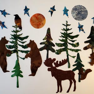 May include: A whimsical forest scene with brown bears, a moose, and squirrels. The image includes green trees, a raven, a sun, a moon, and blue star shapes. The cut-out shapes are arranged on a white background.