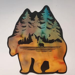 May include: A bear-shaped cutout featuring a colorful watercolor-style background in shades of orange, yellow, and teal. The silhouette of a forest with pine trees, mountains, a crescent moon, and a small bear standing near water is overlaid in black.