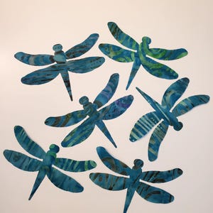 May include: Six dragonfly cutouts featuring a batik-style pattern in shades of blue, teal, and green. Each insect shape displays unique abstract markings, including stripes, dots, and swirls, rendered in a flat, two-dimensional design.