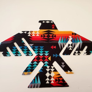May include: A decorative textile in the shape of a stylized bird with outstretched wings. The bird features a geometric pattern in shades of black, red, orange, blue, and gray. The design is set against a plain, off-white background.