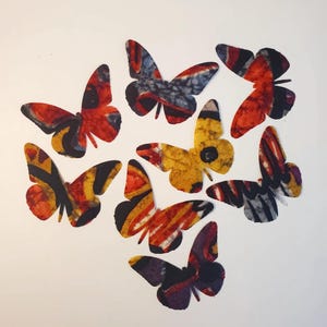 May include: A collection of seven butterfly-shaped cutouts featuring batik-style patterns in vibrant colors including red, orange, yellow, black, and purple. Each butterfly displays unique abstract designs, textured markings, and varied wing shapes.