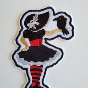 May include: Embroidered iron-on patch featuring a black and white figure of a girl wearing a black dress, red stockings, and a black hat with a white bow. She is holding a black bird in her right hand.