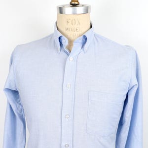 Può includere: Camicia azzurra con bottoni, colletto button-down e taschino sul petto. La camicia a maniche lunghe è esposta su un manichino. La camicia ha un design classico ed è realizzata in un tessuto morbido e tessuto.