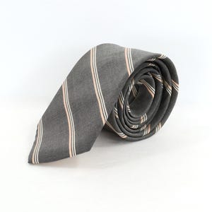 Puede incluir: Una corbata gris enrollada con rayas verticales en rosa claro y blanco. La corbata está hecha de una tela texturizada y se muestra sobre un fondo blanco. Un accesorio clásico para vestimenta formal o de negocios.