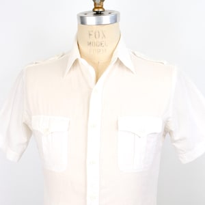 Puede incluir: Camisa de manga corta color blanco roto con dos bolsillos en el pecho y cierre de botones. La camisa se exhibe en un maniquí. El cuello es puntiagudo y las mangas son cortas.