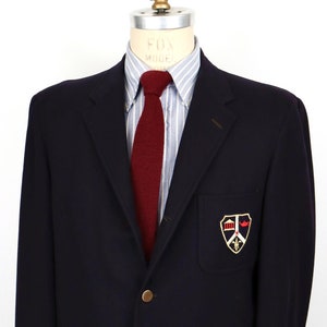 May include: A navy blue blazer with a gold button closure and a patch on the left breast featuring a crest with a red and gold design.