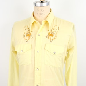 May include: A yellow long-sleeved western shirt with a button-down collar and two chest pockets. The shirt features embroidered horseshoes and horses on the front.