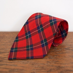 May include: A red plaid necktie with a classic pattern. The tie features a bold red base color with intersecting lines in navy blue, green, and white. The tie is folded and rests on a wooden surface.