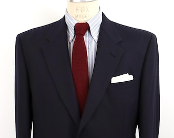 May include: A navy blue blazer with a single gold-tone button closure, worn over a light blue and white striped button-down shirt. A solid dark red knit necktie and a white pocket square are tucked into the blazer pocket on the left side.
